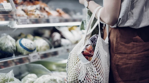 Henkilö, jolla on yllään hame ja t-paita kantaa käsivarrellaan narukassia, jossa on juureksia ja vihanneksi, ja katsoo kaupan hyllyllä olevia juureksia.