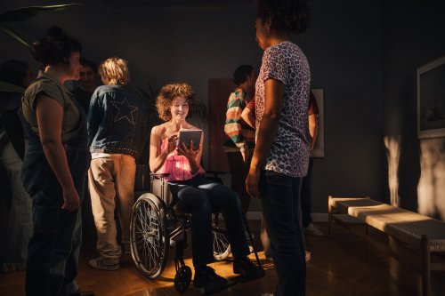 Useita henkilöitä huoneessa, henkilö keskellä katsoo tablettitietokonetta istuen pyörätuolissa