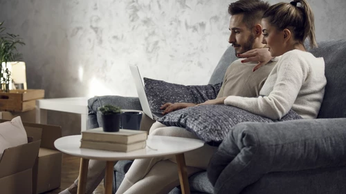 Nuorehko nainen ja mies istuvat harmaalla sohvalla ja katlevat yhdessä miehen sylissä olevaa läppäriä. Huoneessa on muuttolaatikoita.