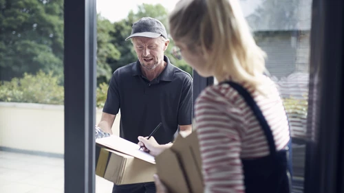 Mies lippalakissaan toimittajaa paketin ulko-ovella seisovalle naiselle.