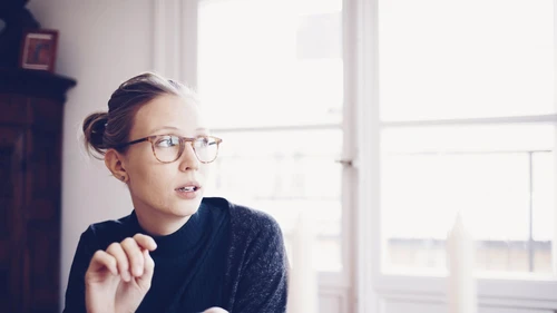 Nuori silmälasipäinen nainen katsoo huolestuneena ikkunasta ulos puhelin kädessään.