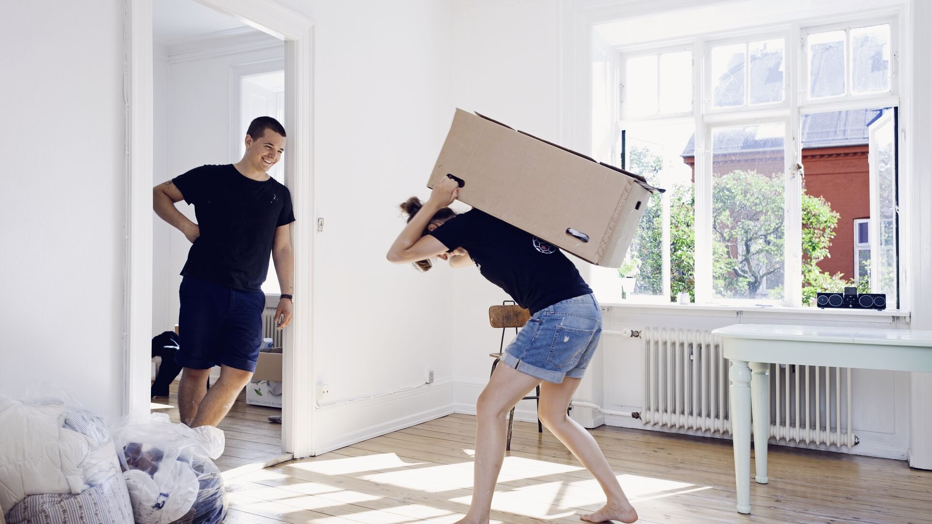 Man watching girlfriend carry heavy box in apartment