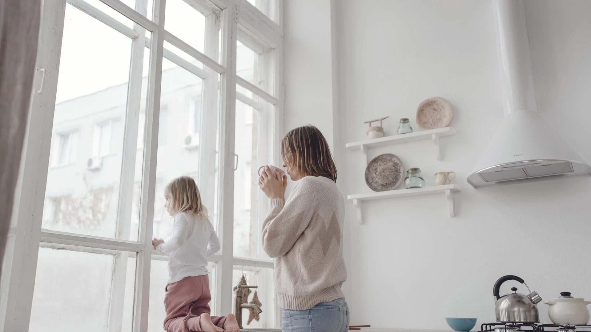 Nainen ja tyttö katsovat ulos isosta ikkunasta vaaleansävyisessä huoneessa.