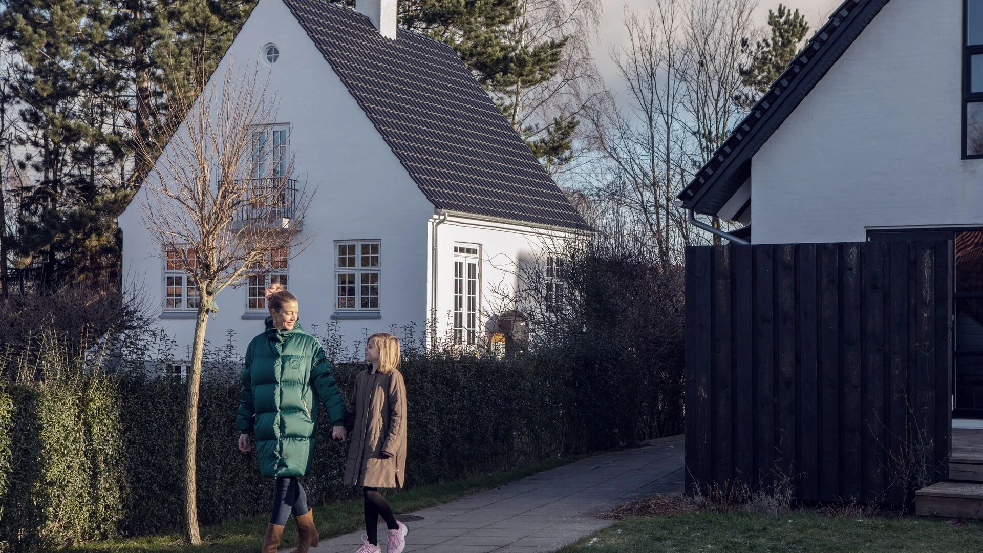 Talvivaatteisiin pukeutuneet äiti ja tytär kävelevät valkoisten omakotitalojen edessä toisiinsa katsoen.