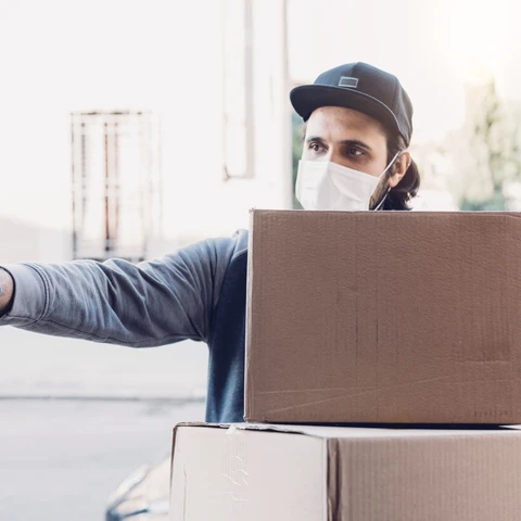 Delivery man is ringing the bell to deliver two cardboard boxes. He's wearing a face mask and protective gloves.