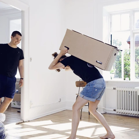 Man watching girlfriend carry heavy box in apartment