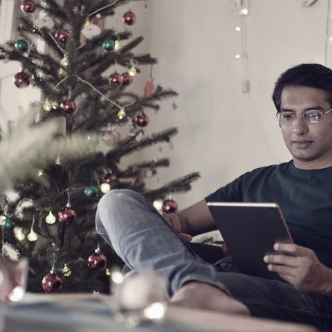 man sitting next to a christmas tree reading on his ipad