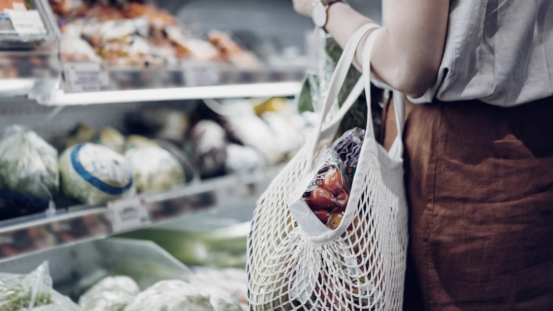 Henkilö, jolla on yllään hame ja t-paita kantaa käsivarrellaan narukassia, jossa on juureksia ja vihanneksi, ja katsoo kaupan hyllyllä olevia juureksia.