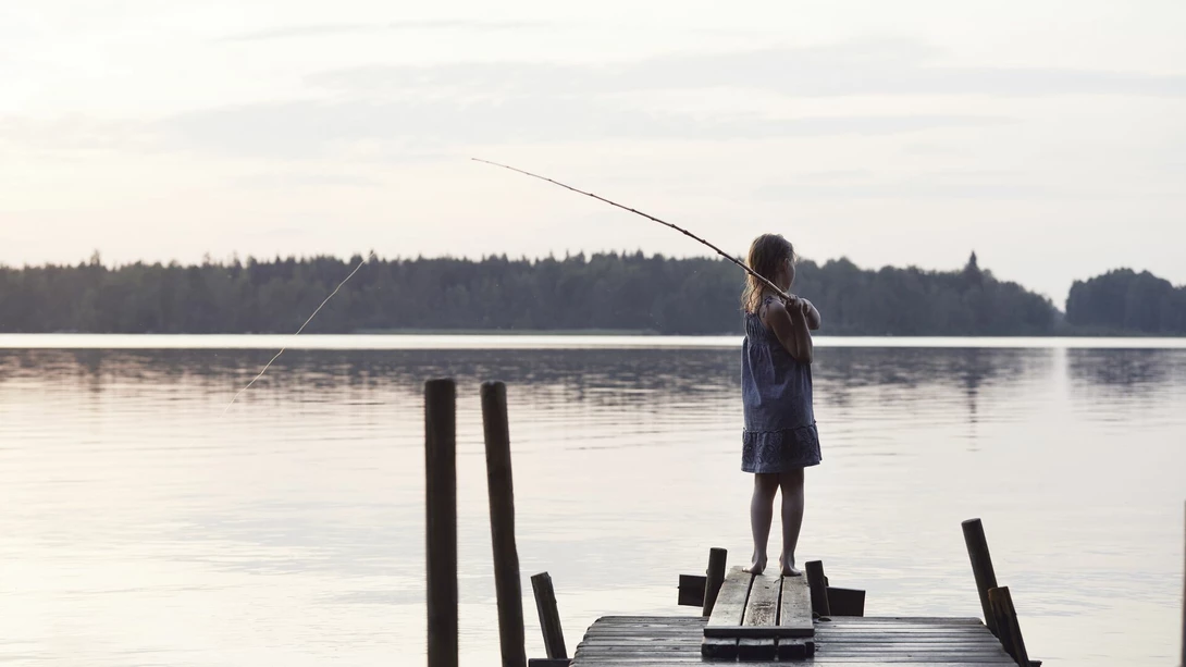 Pieni tyttö sinisessä kesämekossa onkii puisella laiturilla. Järvi on tyyni ja taustalla näkyy havupuumetsää.